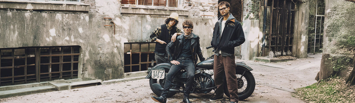 Tres modelos usando prendas workwear, cuero y layering junto a motocicleta custom en entorno urbano desgastado — FALL/WINTER 2026 by DEMONS & MACHINES