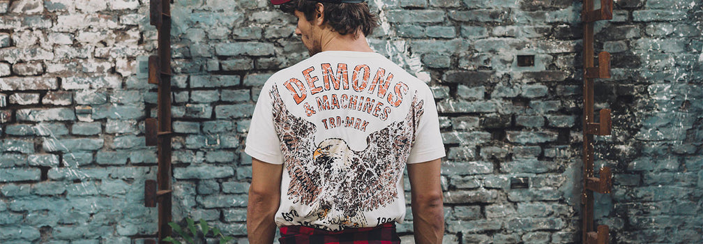 Remera Demons & Machines con estampa gráfica trasera de águila, estilo vintage, en look masculino urbano frente a muro de ladrillo