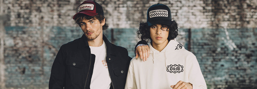 Dos modelos con indumentaria Demons & Machines, gorras y hoodie en look urbano frente a muro de ladrillo