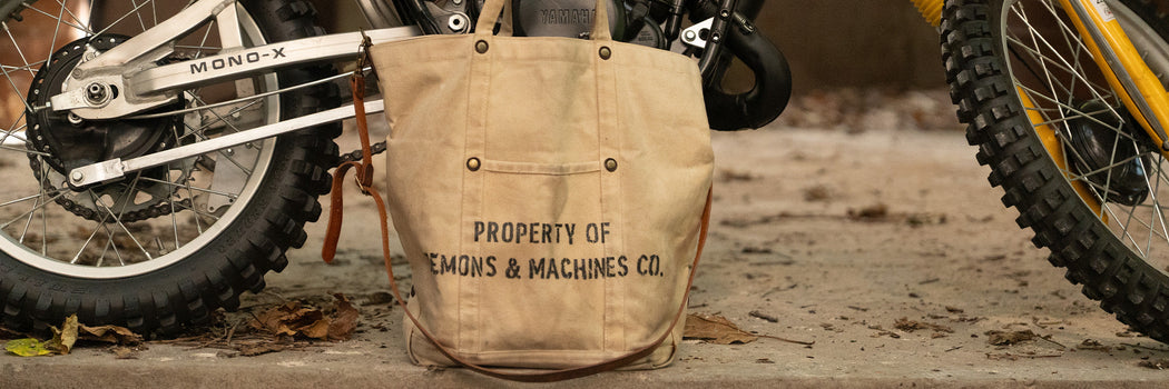 BOLSO TOTE DE LONA DEMONS & MACHINES CON ESTAMPA “PROPERTY OF” APOYADO ENTRE MOTOS OFF-ROAD