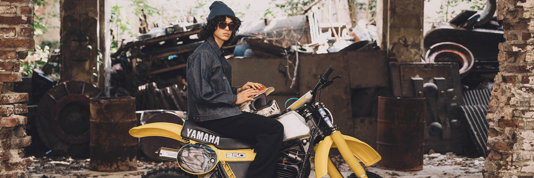 MODELO CON CAMPERA Y HOODIE NEGRO DEMONS & MACHINES SOBRE MOTO YAMAHA AMARILLA EN ESCENA INDUSTRIAL