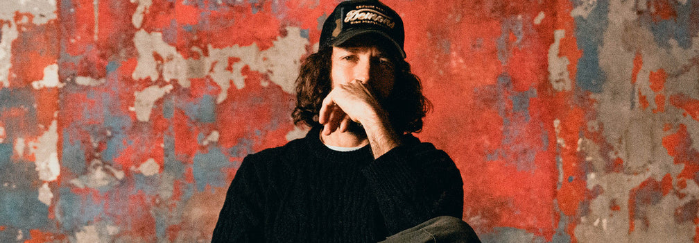 Modelo usando gorra trucker y prendas oscuras frente a muro desgastado con iluminación cálida industrial — FALL/WINTER 2026 by DEMONS & MACHINES