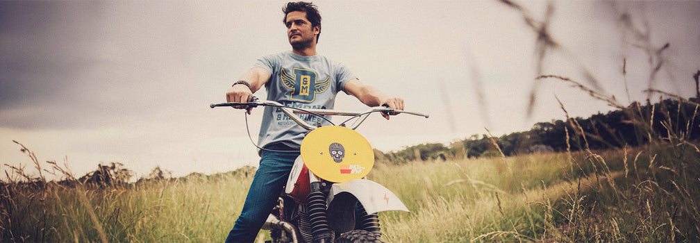 HOMBRE SOBRE MOTO OFF-ROAD EN CAMPO ABIERTO CON REMERA DEMONS & MACHINES Y ESTÉTICA VINTAGE