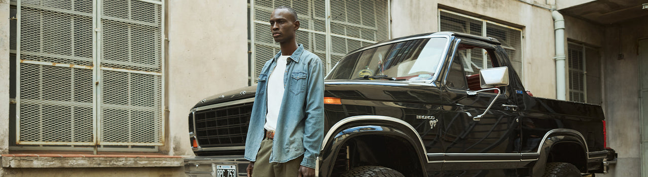 Hombre con camisa de jean junto a una pickup negra antigua frente a un edificio — Drop 1 SS27