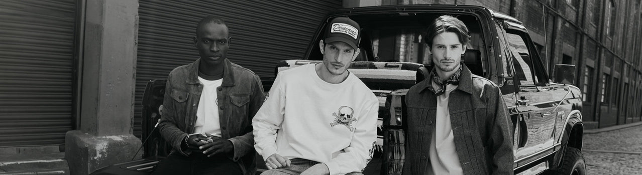 Tres hombres con remera blanca, buzo blanco estampado y camisa junto a una camioneta antigua, en blanco y negro — SS27