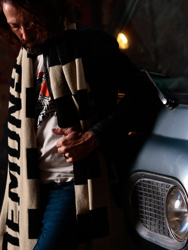 Modelo usando bufanda oversized y remera gráfica junto a vehículo vintage Ford Falcon en escena industrial nocturna — DROP 2 FW26 by DEMONS & MACHINES
