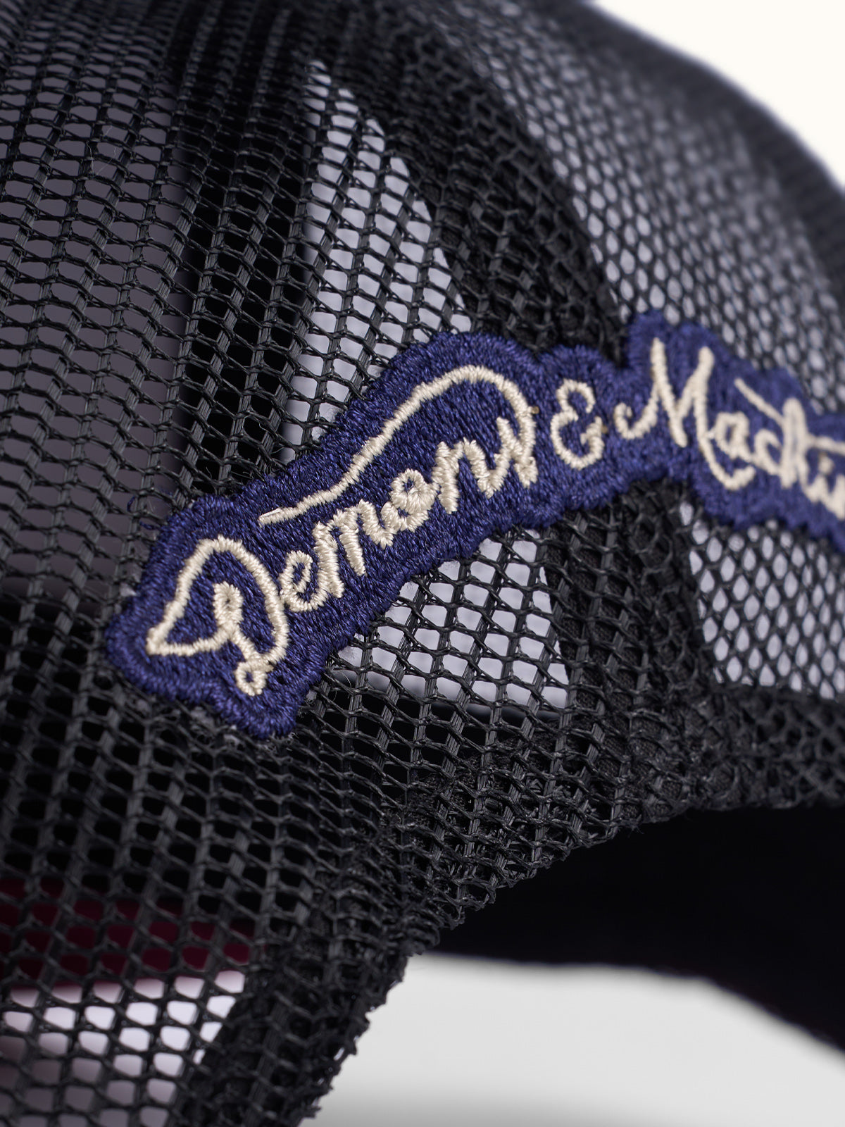 Primer plano del bordado Demons & Machines en cursiva sobre la red trasera de la Gorra Trucker bordo