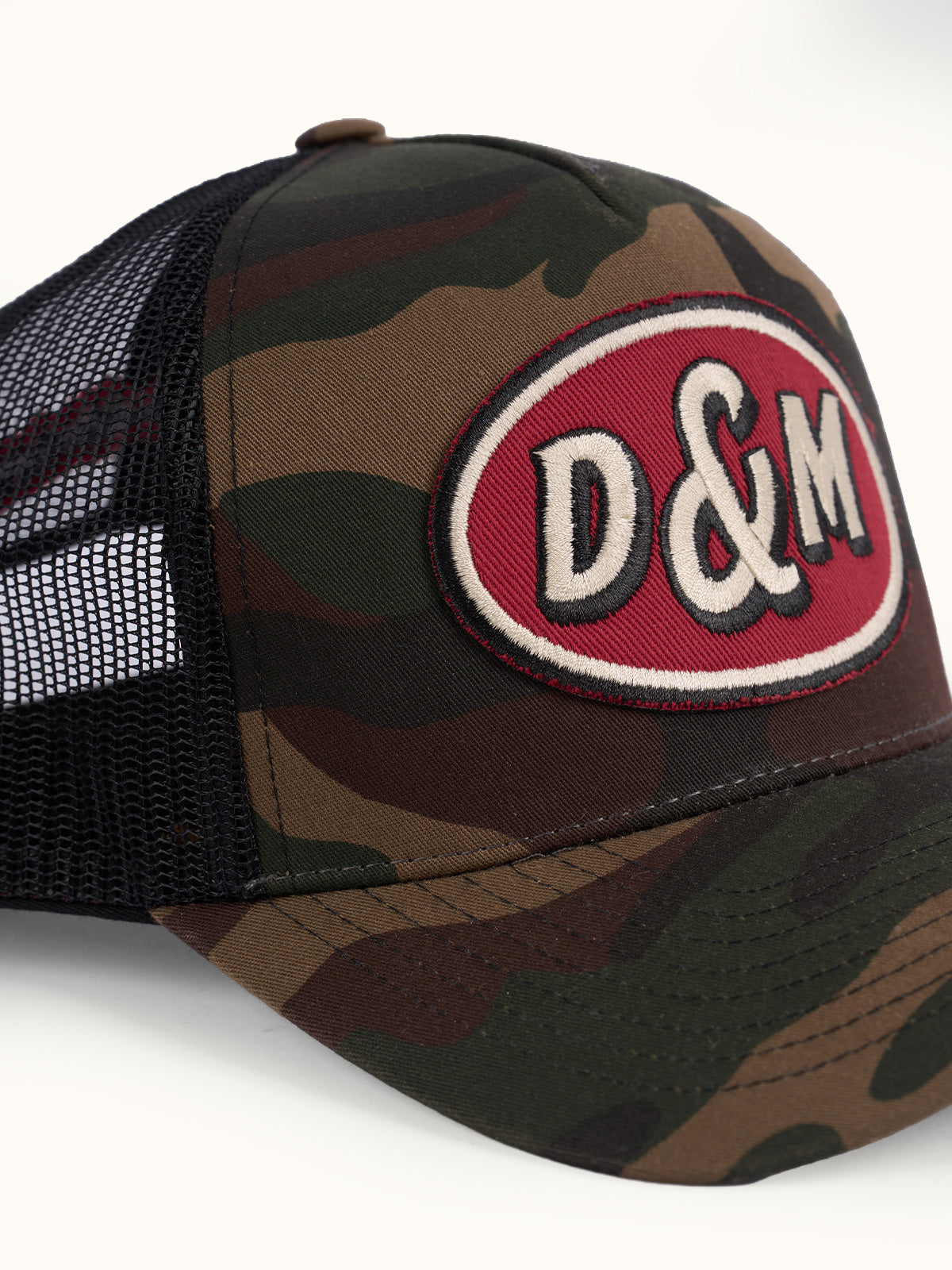 Primer plano del parche ovalado rojo D&M bordado al frente de la Gorra Trucker camuflada