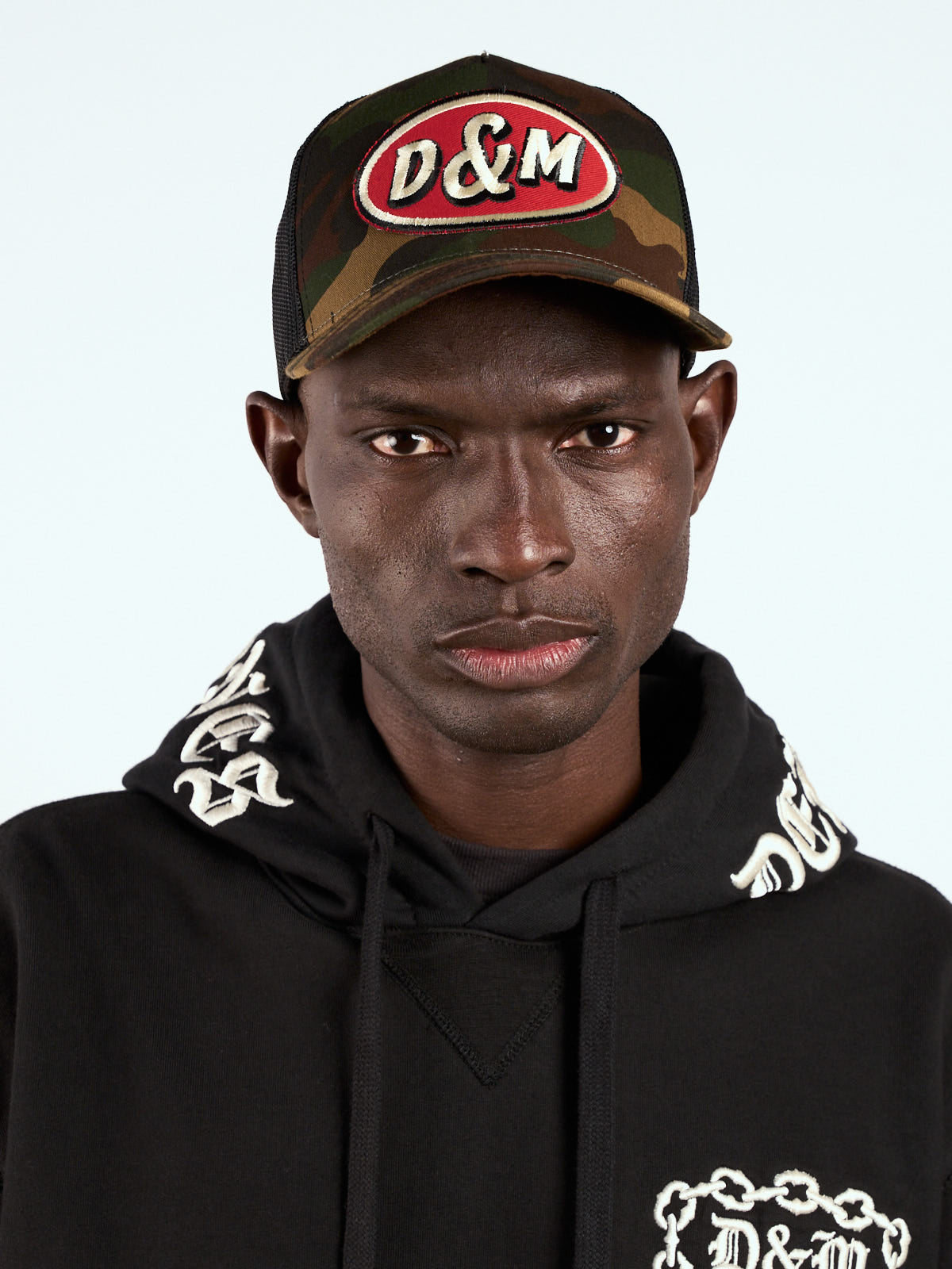 Gorra Trucker camuflada puesta, retrato de frente con parche D&M y hoodie negro bordado #color_camuflado