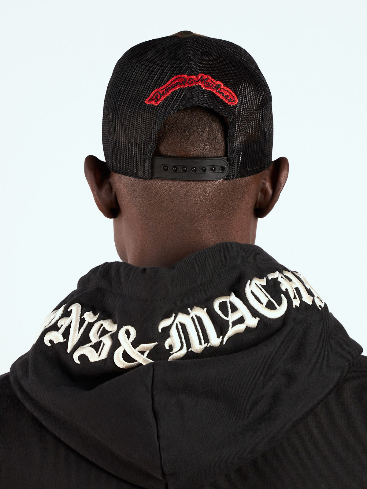 Gorra Trucker camuflada puesta, vista de espalda con bordado script rojo sobre la red y hoodie negro