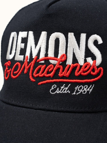 Primer plano del bordado Demons en blanco y Machines en cursiva roja de la Gorra Trucker D&M #color_negro