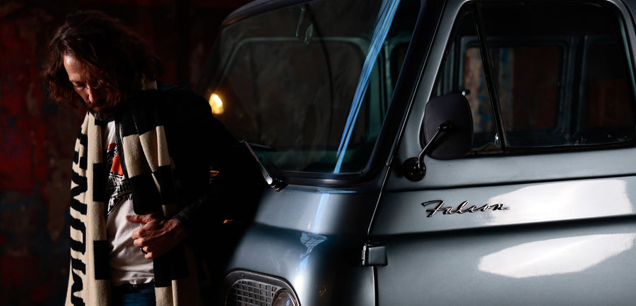 Modelo usando bufanda oversized y remera gráfica junto a vehículo vintage Ford Falcon en escena industrial nocturna — DROP 2 FW26 by DEMONS & MACHINES