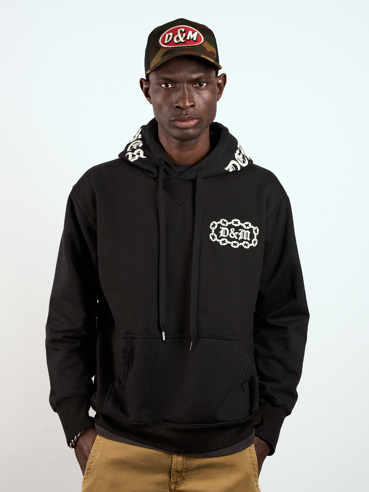 Hoodie Bordado Chain negro puesto, de frente con bordado de cadena en el pecho y gorra trucker #color_negro