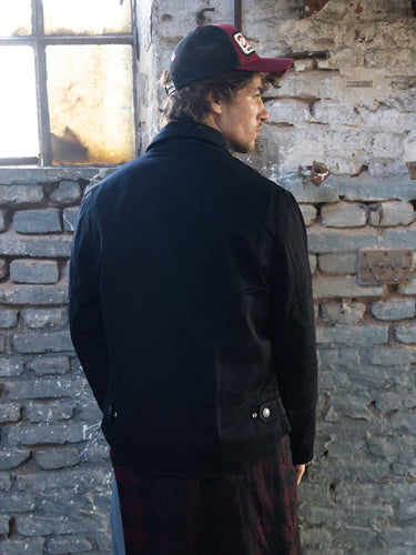 Campera Roswell Cotton negra puesta, vista de espalda con gorra bordó ante pared de ladrillo y ventana #color_negro