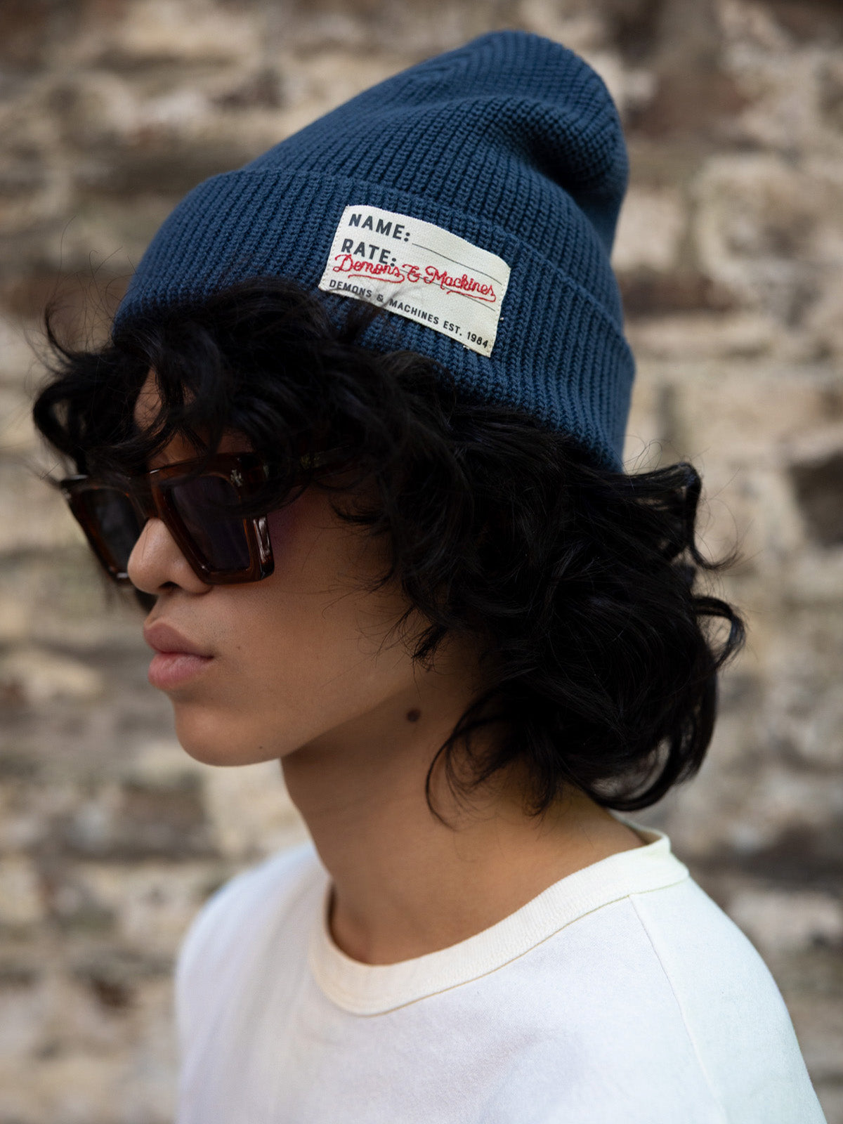 Beanie Tejido azul puesto, de perfil con anteojos oscuros, remera blanca y pared de piedra #color_azul