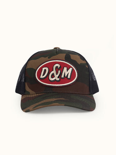 Gorra Trucker camuflada de frente con parche ovalado D&M rojo y crudo y red trasera negra #color_camuflado