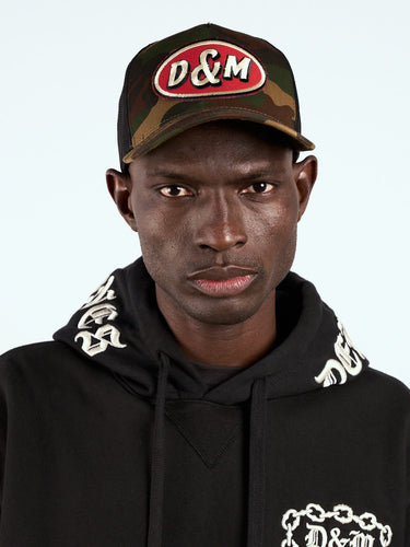 Gorra Trucker camuflada puesta, retrato de frente con parche D&M y hoodie negro bordado #color_camuflado