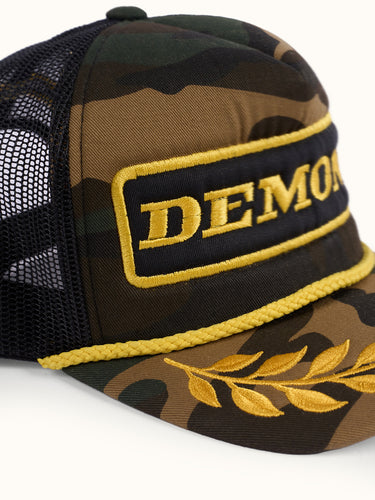 Detalle en tres cuartos de la Gorra Podium II camuflada con parche DEMONS, cordón dorado y laureles #color_camuflado