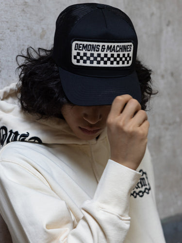 Gorra Trucker II negra puesta, modelo con buzo crudo tocando la visera ante pared de hormigón #color_negro