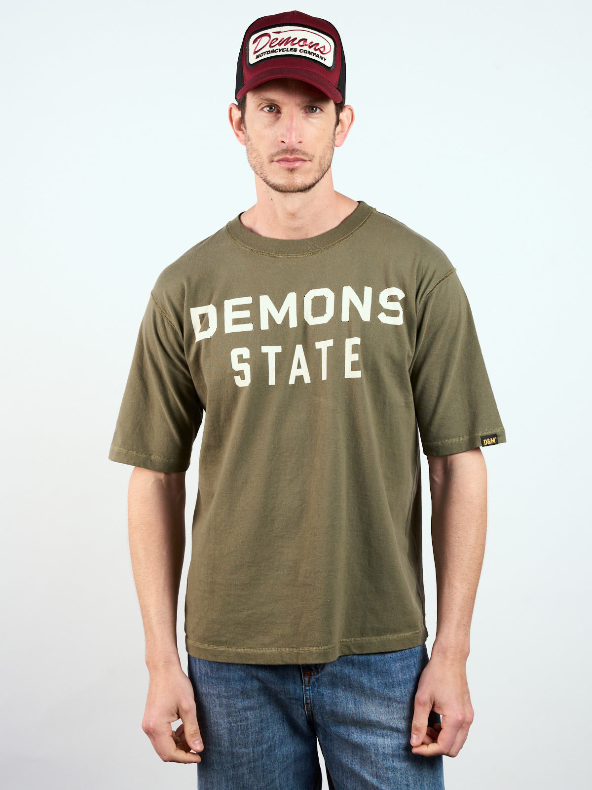 Remera Relax State verde puesta, de frente con estampa blanca Demons State, gorra trucker y jean #color_verde